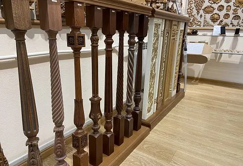 Фото деревянной балюстрады и колонн с разнообразными резными элементами и декоративными панелями, расположенных в выставочном зале с деревянным полом и стенами, украшенными орнаментами.