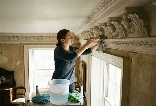 Женщина аккуратно очищает рельефный потолочный карниз, используя кисть и ткань; рядом стоит ёмкость с водой, интерьер выглядит старинным, с богатой декоративной лепниной.