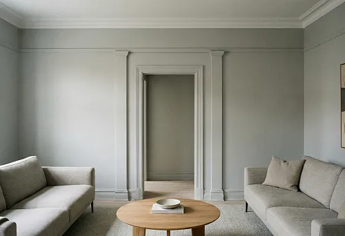 Светлая гостиная с лепным декором: дверной портал с пилястрами и карнизом на серых стенах, два дивана и круглый деревянный стол.